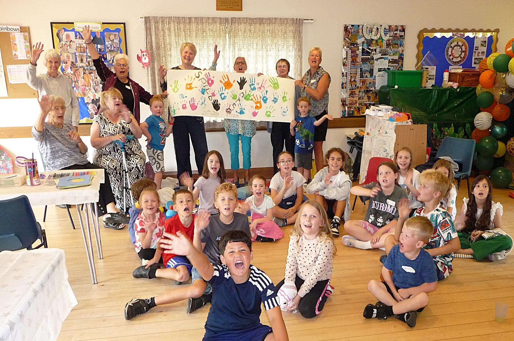 Group of children sat on the floor, happily smiling with adults behind them happily holding up a "Spud Club" handpainted sign